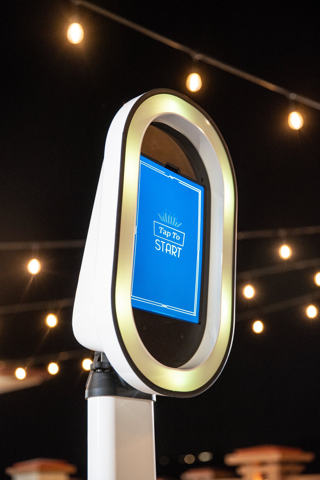 White photo booth with a glowing ring and a blue Tap To Start screen under string lights at night