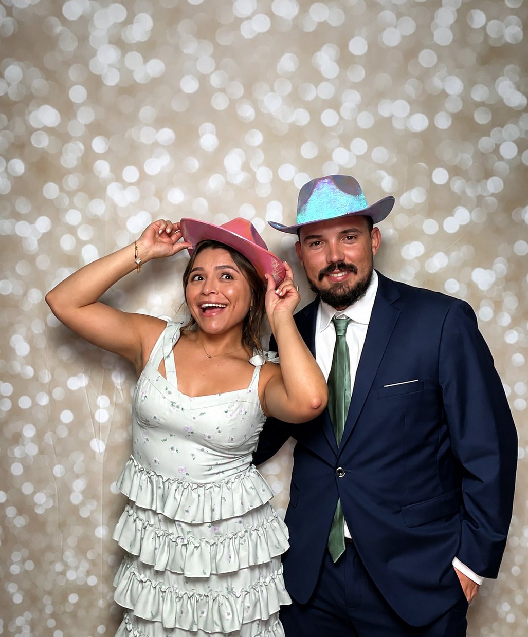A couple in cowboy hat props posing against a champagne sparkle backdrop, she in a ruffled dress