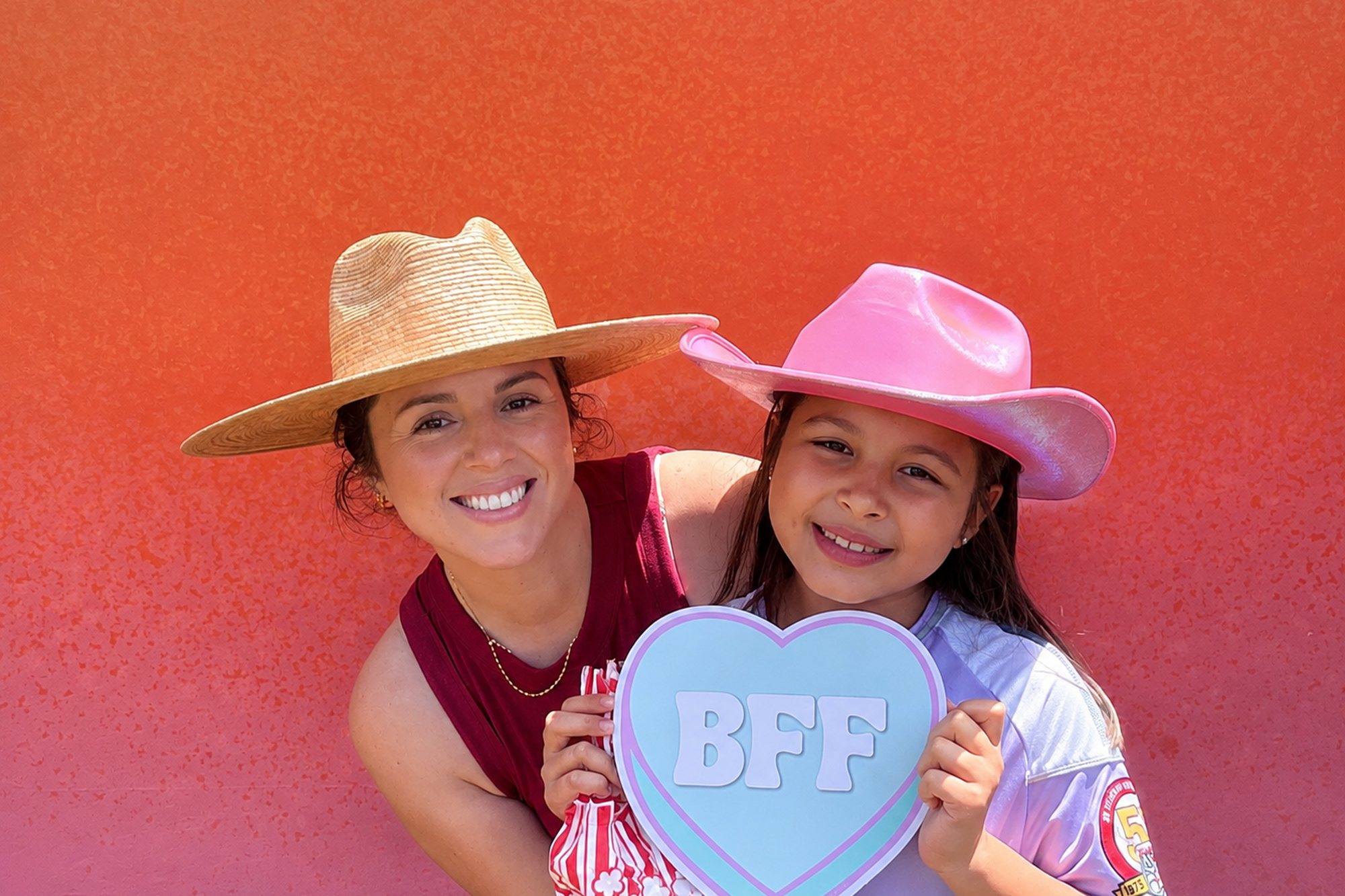 Two guests in felt hats holding a BFF heart prop against a warm color gradient