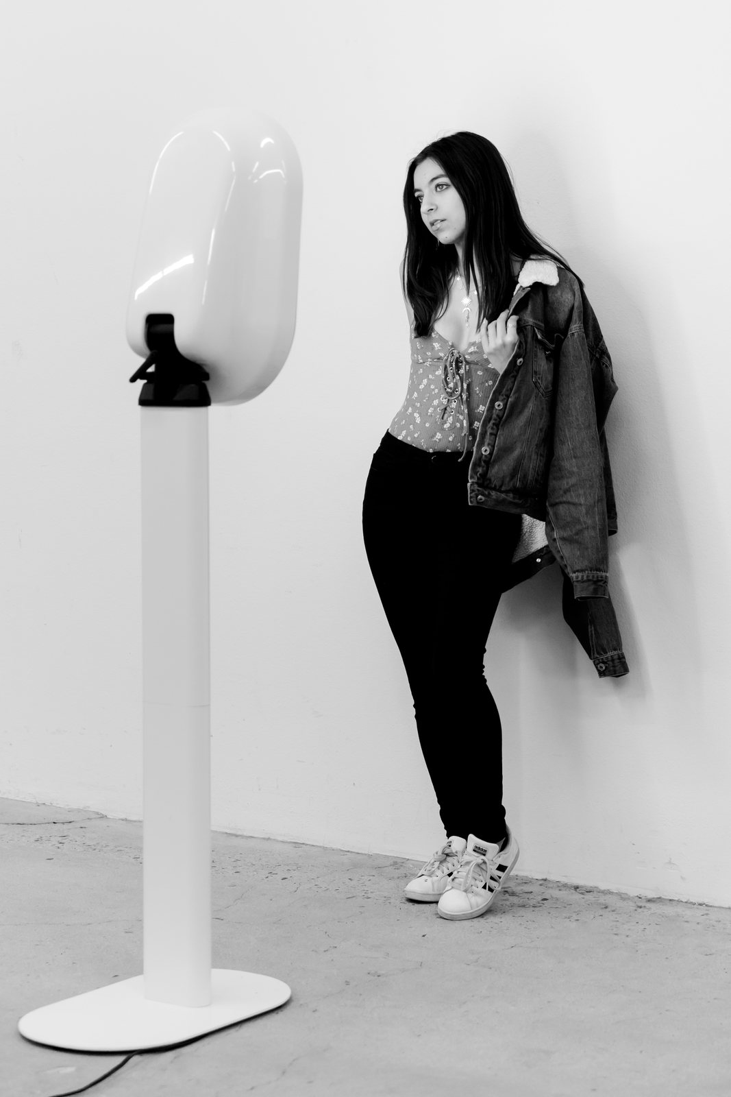 Black-and-white shot of a woman in a denim jacket leaning on a wall beside a white photo booth