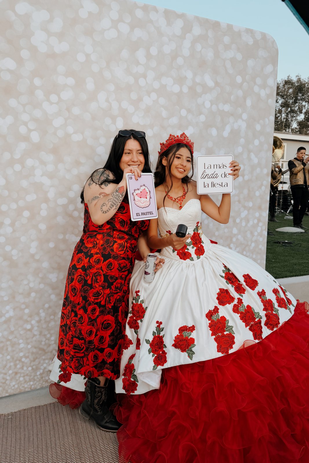 A guest in a red floral gown posing with a fan prop against a glitter backdrop