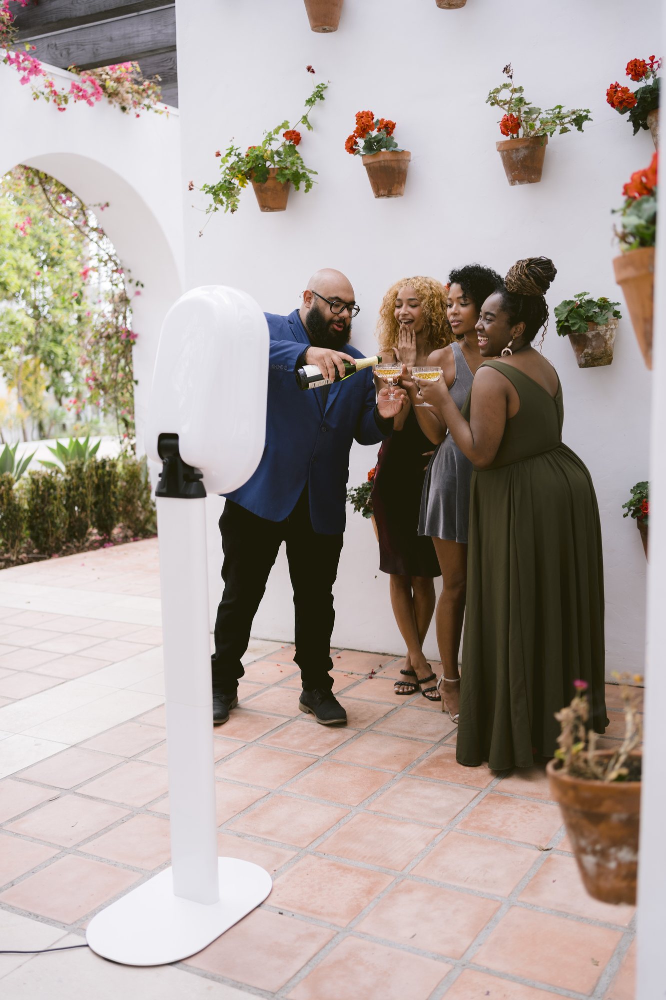 A guest in a blue blazer pouring champagne for three women beside a white photo booth in a courtyard