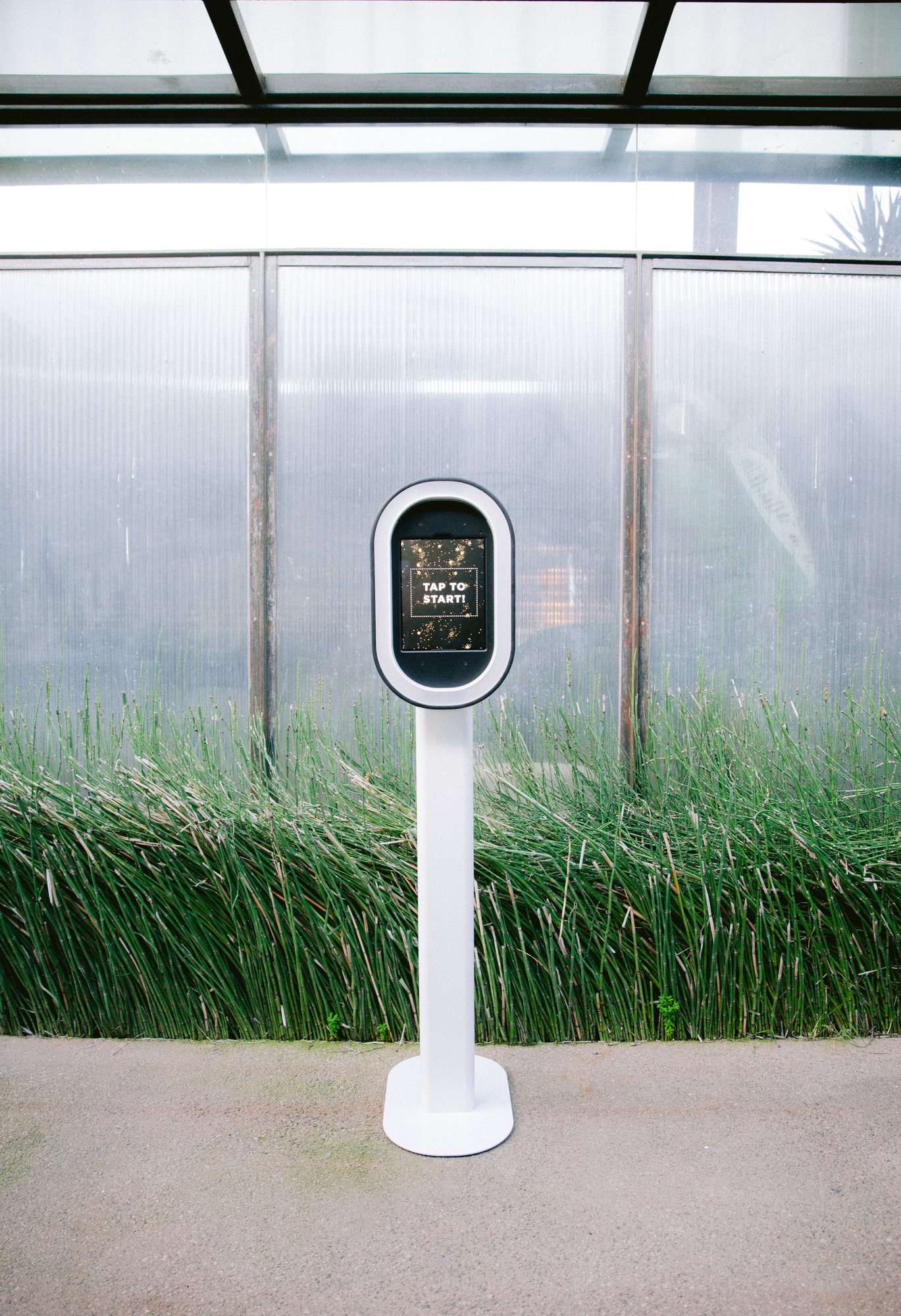 White photo booth with a Tap to Start screen standing before a greenhouse and tall grasses