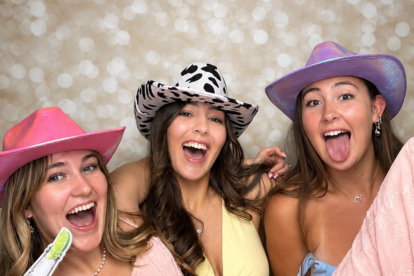 Three guests in party hats laughing together in front of a champagne glitter backdrop